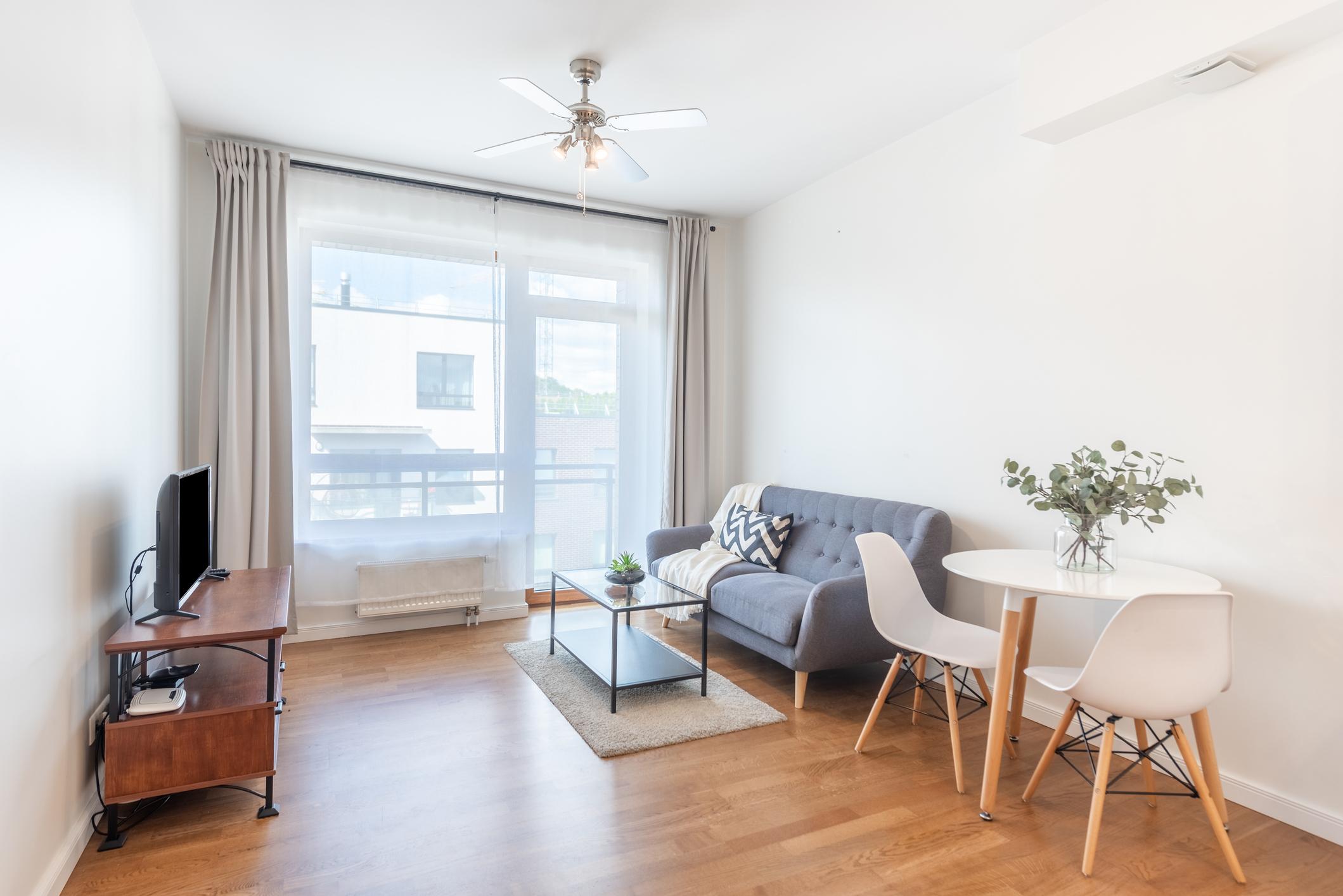 Bright small living room with light colors, minimal furniture, wall shelves, and a mirror creating a spacious and uncluttered look.
